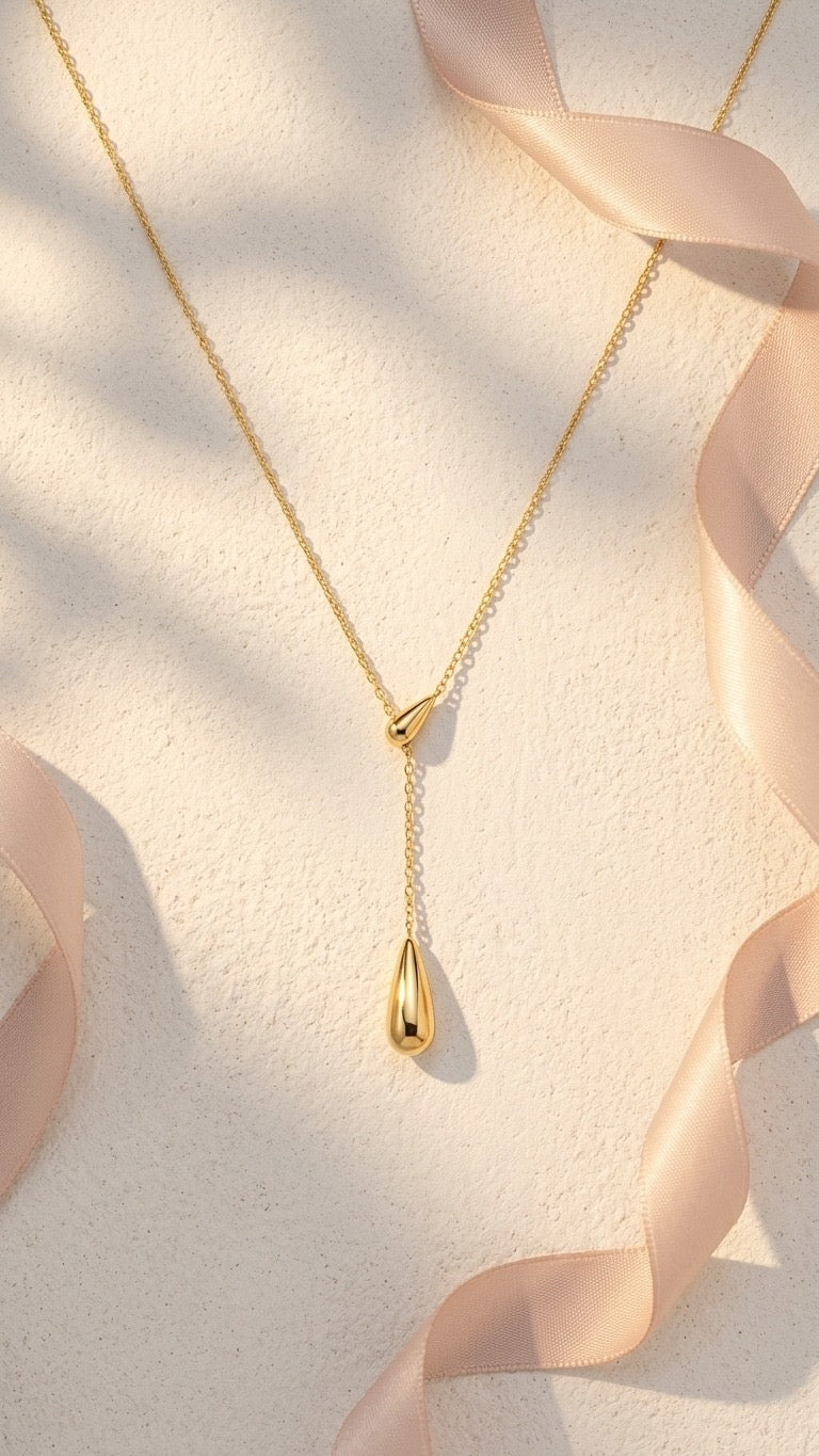 Lilyah Jewelry's Gold necklace with a teardrop pendant on a light background with a white light pink ribbon. The necklace looks like it has two golden drops. It is adjustable, minimal yet a statement piece.